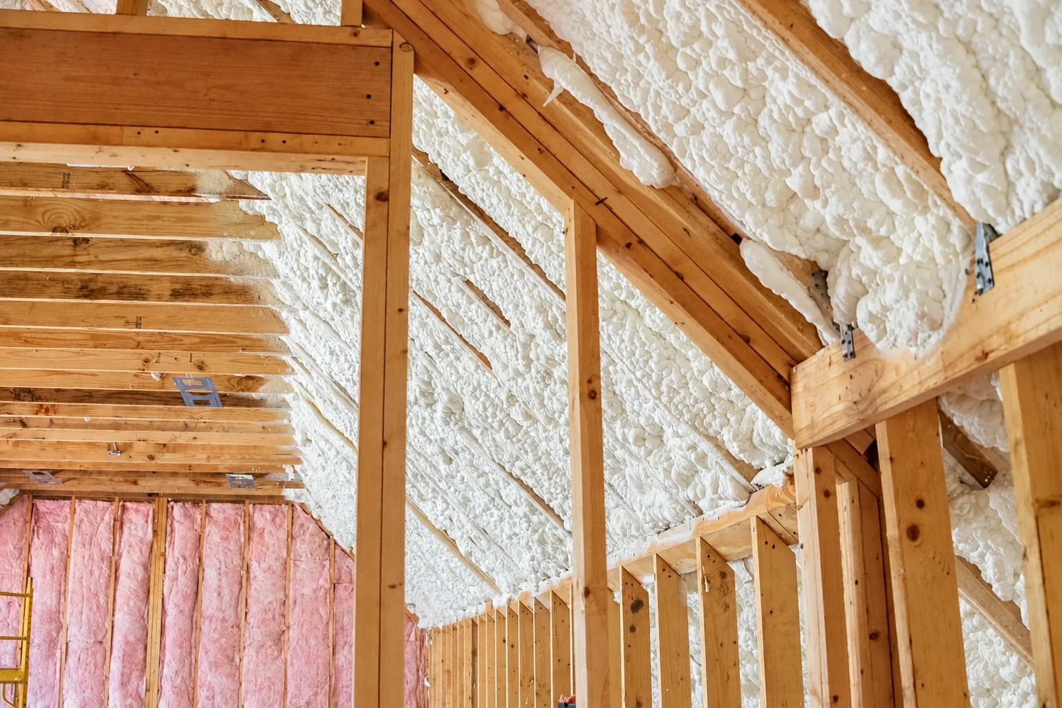 Exposed roof framing coated with white spray foam insulation, with pink batt insulation in the lower wall.