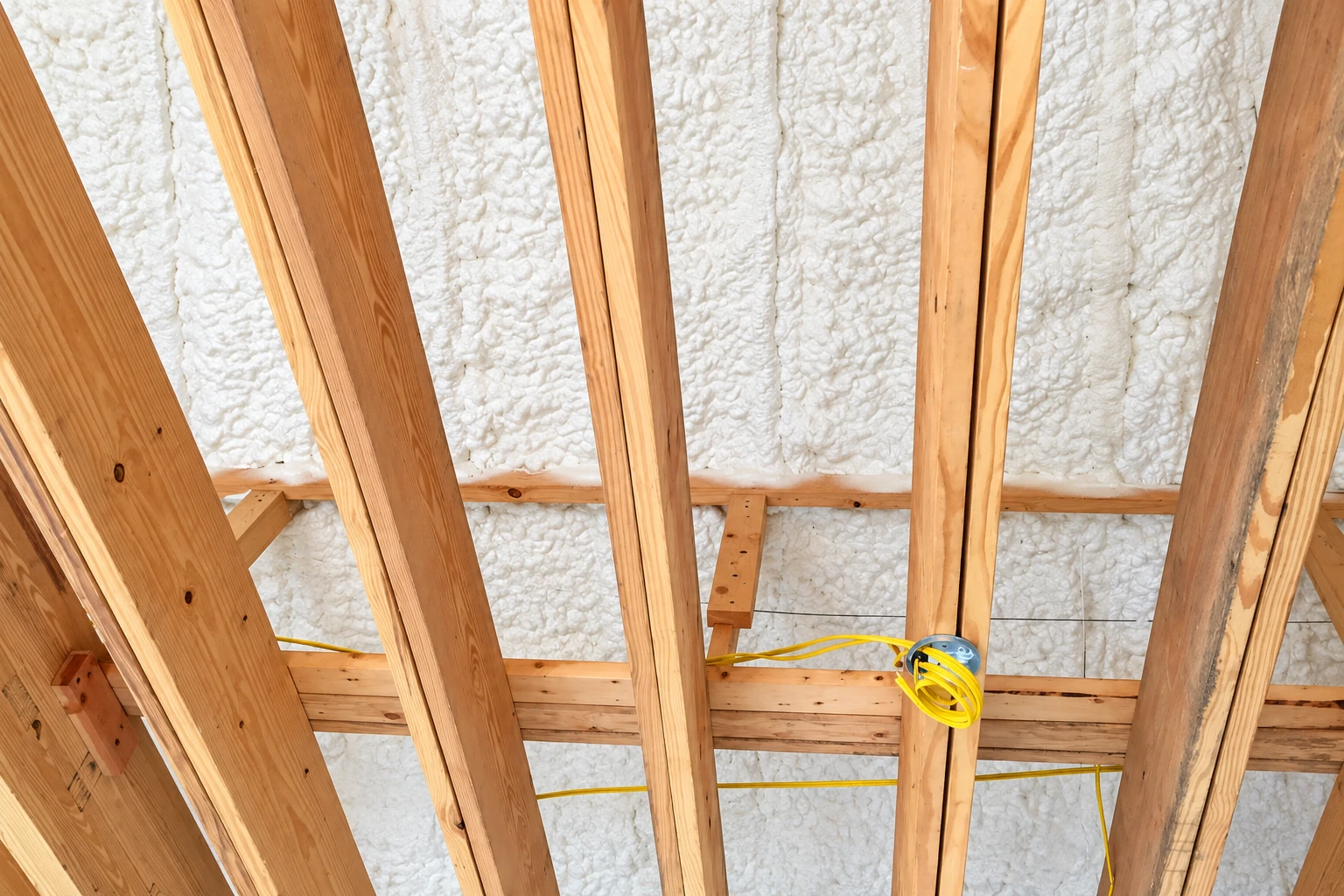 Exposed roof framing with white spray foam filling the spaces between wooden beams and yellow wiring along the framing.