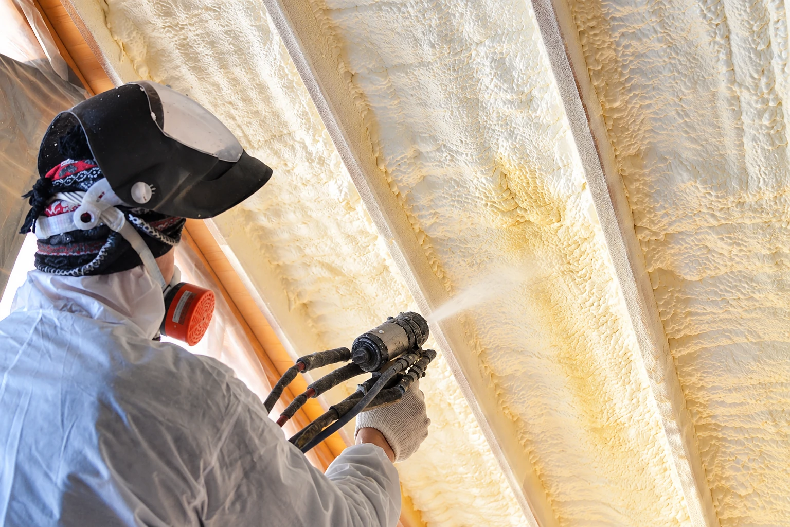 Installer in protective gear sprays closed-cell foam insulation between exposed roof rafters.