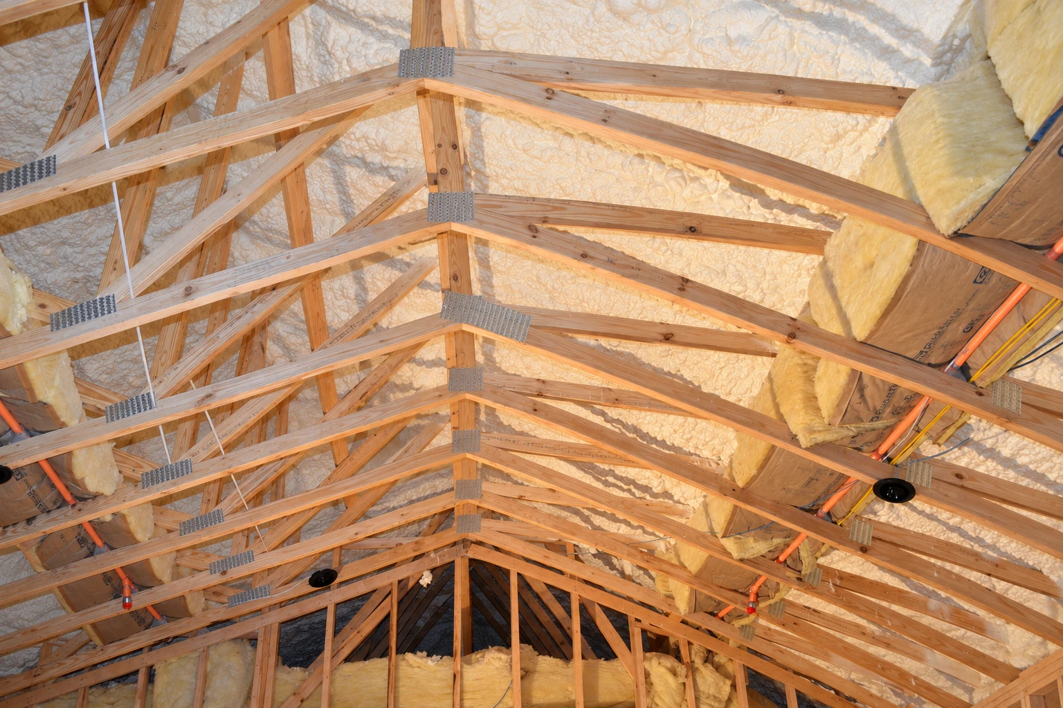 Foam insulation and yellow batts fill the roof cavities between exposed wooden trusses.