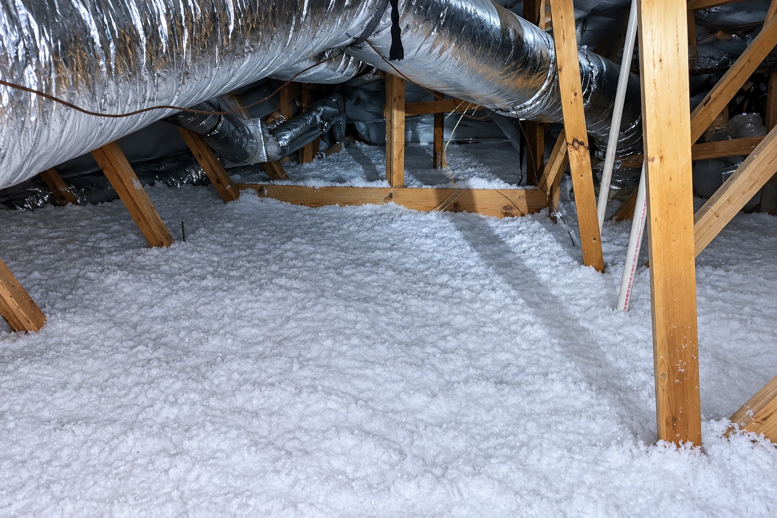An attic floor is covered with loose white insulation beneath foil-wrapped ducts and exposed wooden framing.