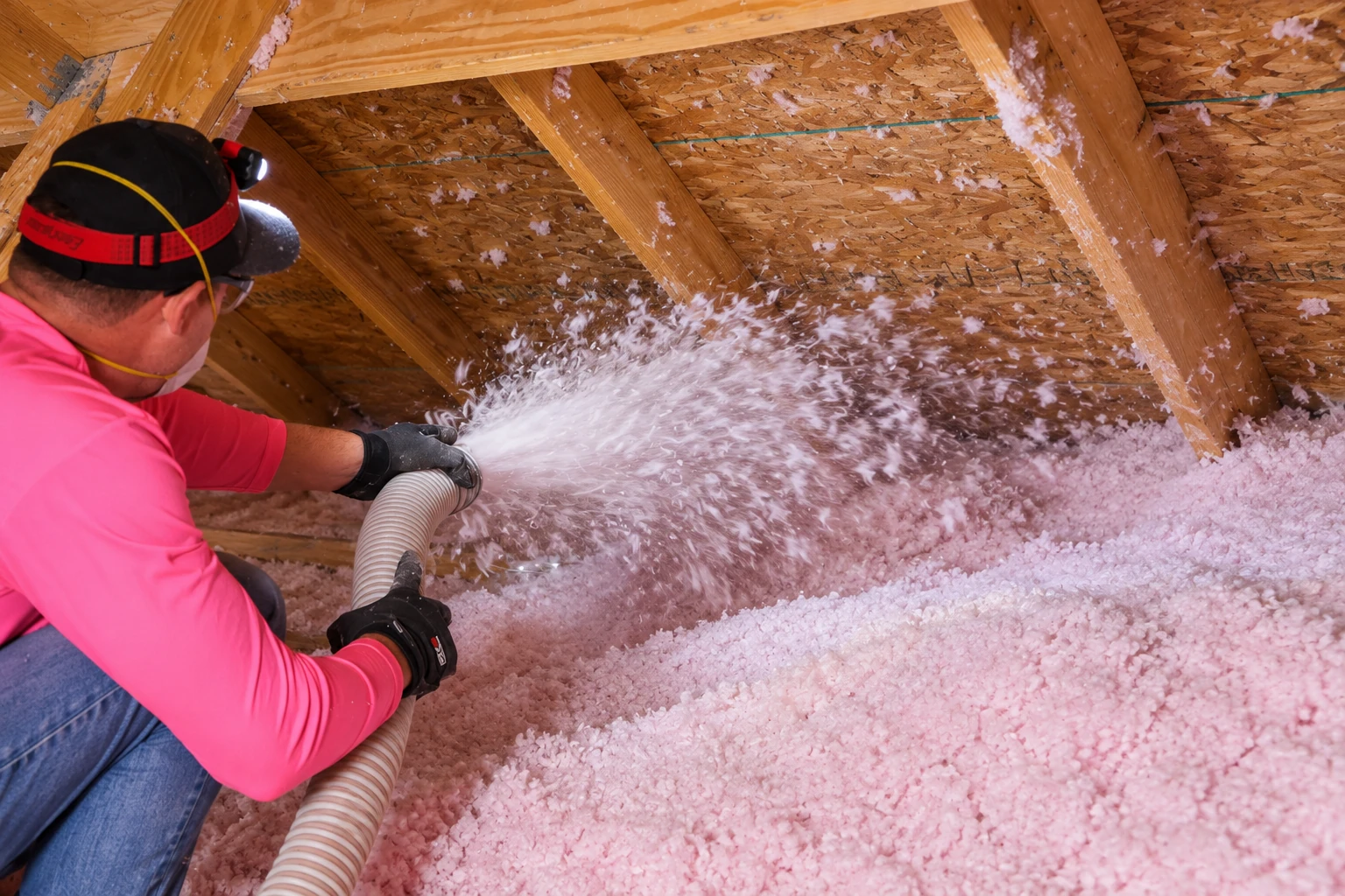 A worker in a pink shirt sprays loose-fill insulation across an attic floor between exposed wooden rafters.