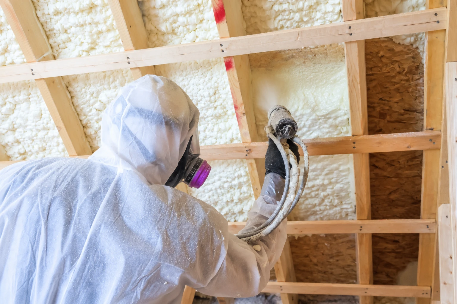 Worker in protective gear applies spray foam insulation to exposed framing with a hose-connected spray gun.
